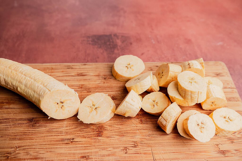 A peeled banana on a wooden cutting board, partially sliced into round pieces like those for patacones, against a pinkish background.