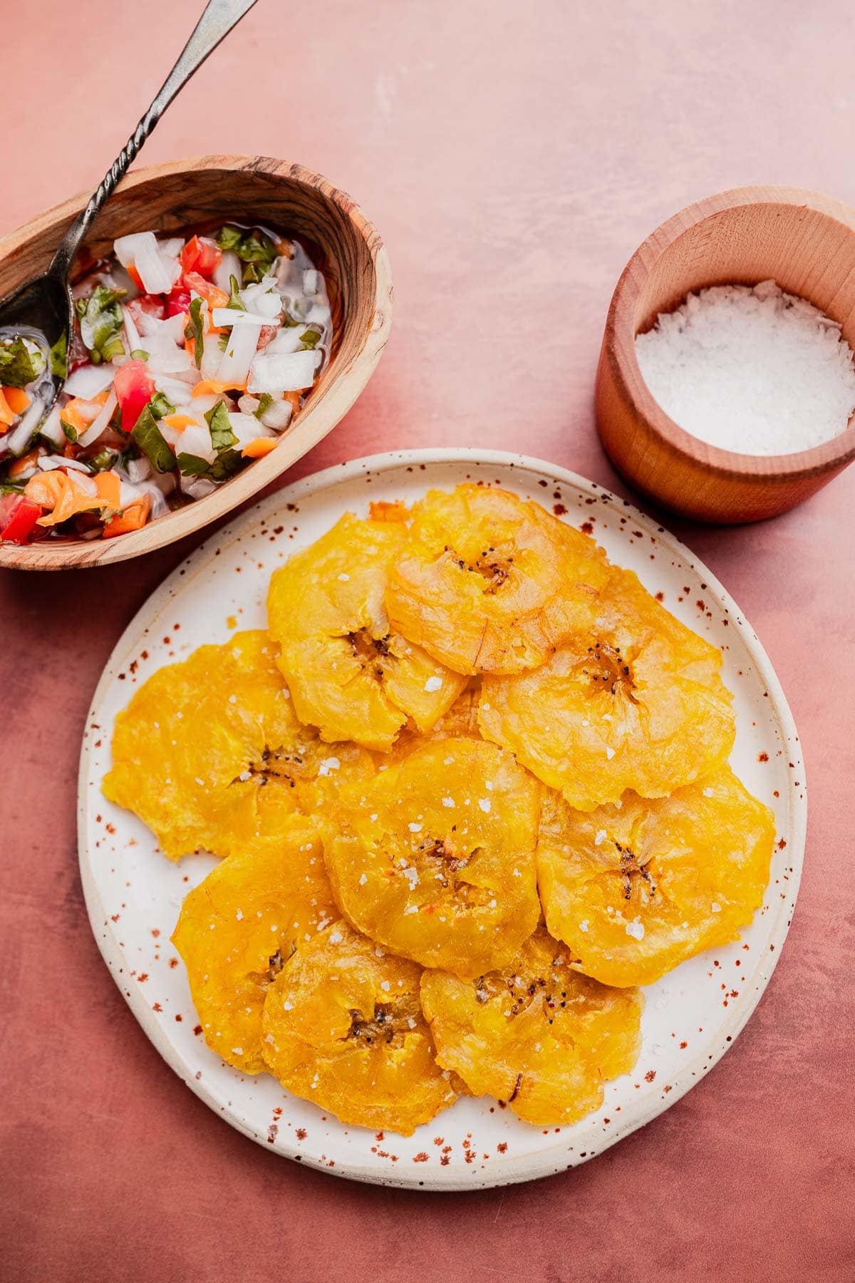 A plate of crispy patacones, or fried plantain tostones, sprinkled with salt sits beside a bowl of chopped vegetable salsa and a small wooden bowl of salt on a pink surface.