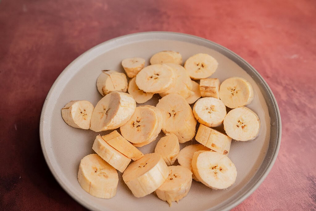 A beige plate holds several slices of raw plantain, ready to be made into patacones, on a reddish-brown surface.