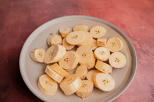 A beige plate holds several slices of raw plantain, ready to be made into patacones, on a reddish-brown surface.