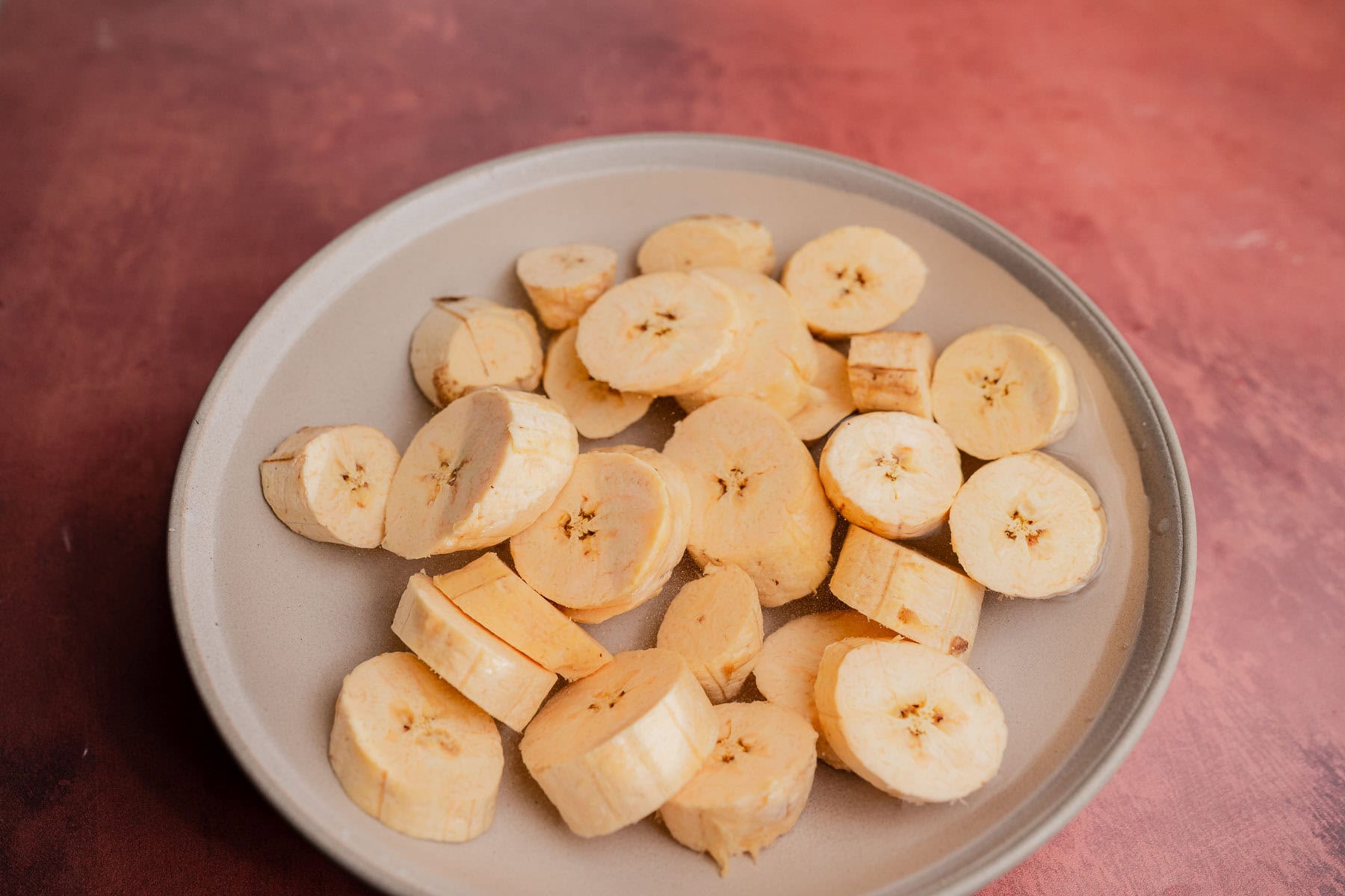 A beige plate holds several slices of raw plantain, ready to be made into patacones, on a reddish-brown surface.
