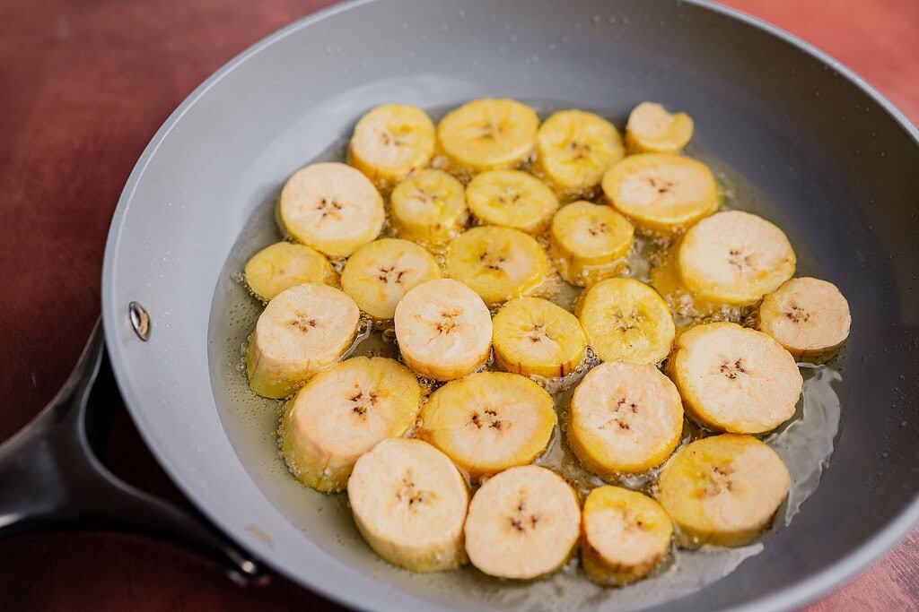 Sliced plantains frying in oil in a non-stick skillet, soon to become crispy patacones.
