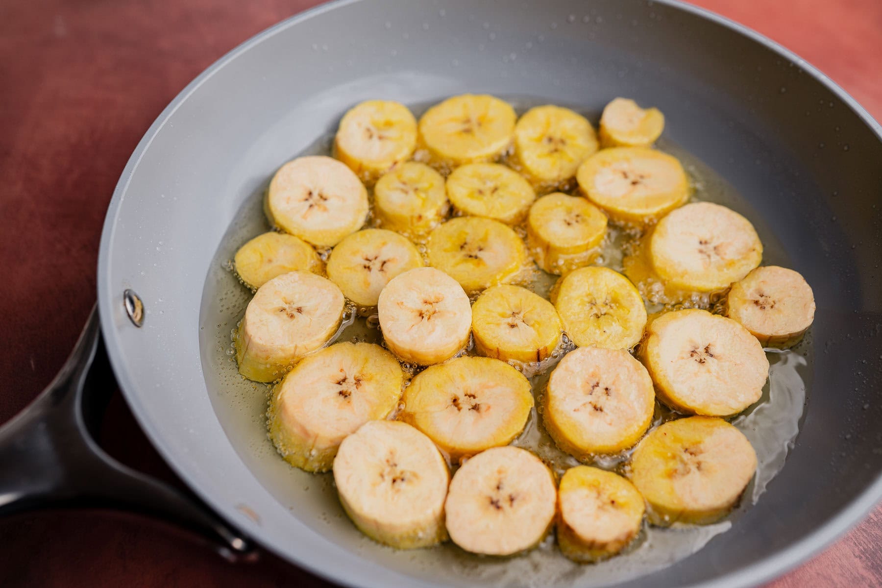 Sliced plantains frying in oil in a non-stick skillet, soon to become crispy patacones.