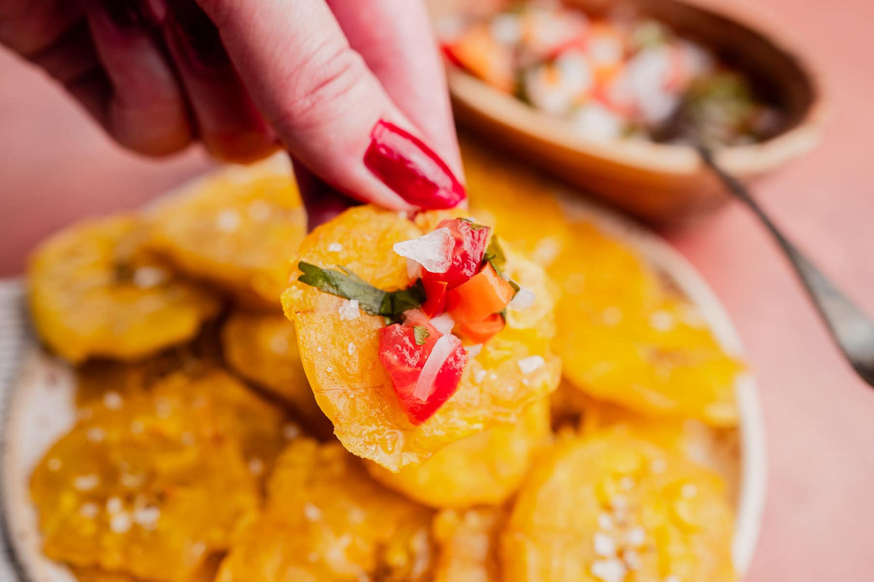 A hand with red nail polish holds a patacón topped with chopped tomatoes, onions, and herbs over a plate full of more tostones.