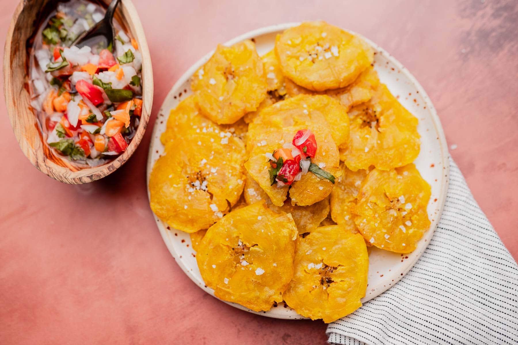A plate of golden fried patacones topped with chopped vegetables and salt, next to a bowl of salsa, on a pink surface with a striped napkin.