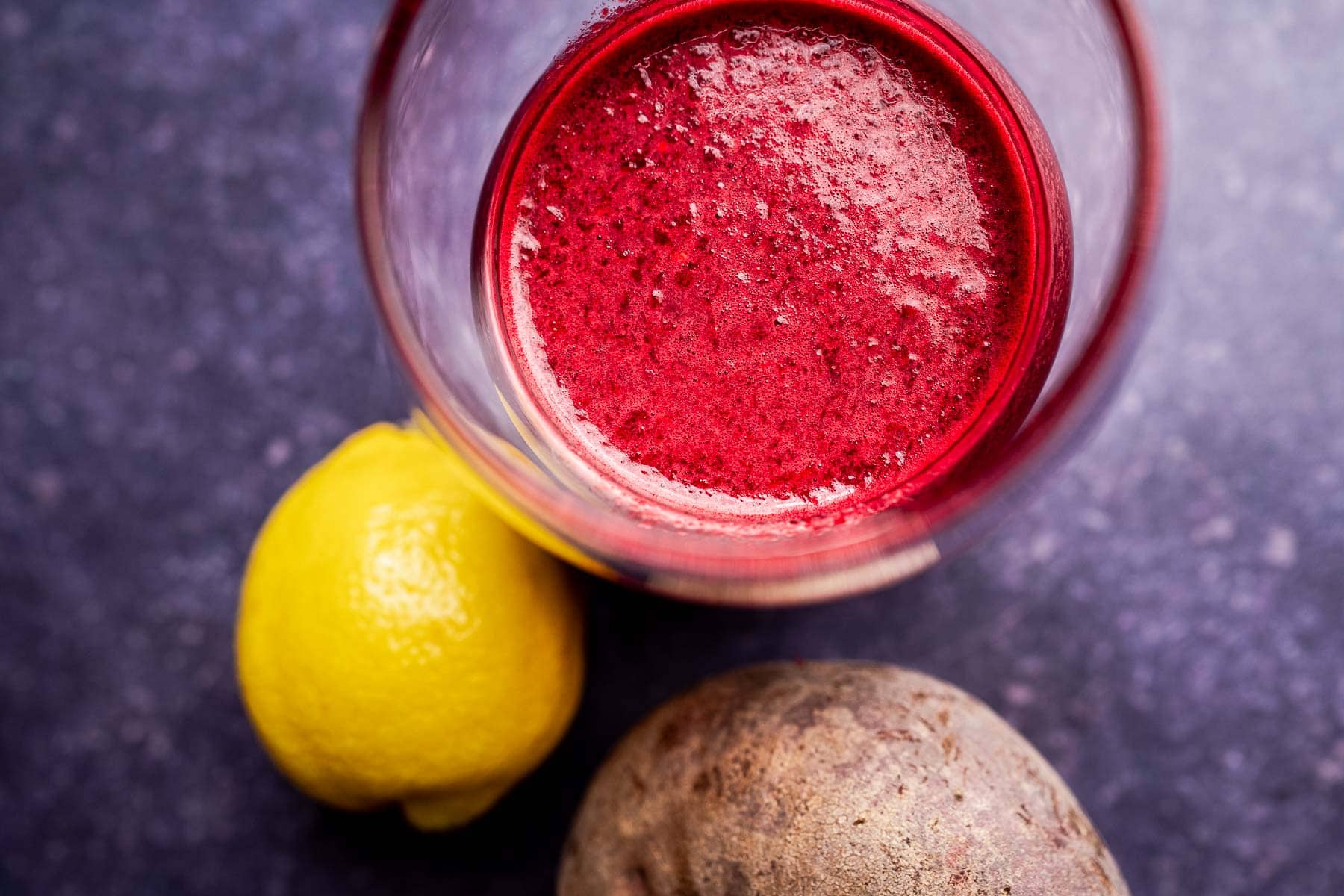 A glass of beet pineapple juice sits beside a whole lemon and a potato on a dark, speckled surface.