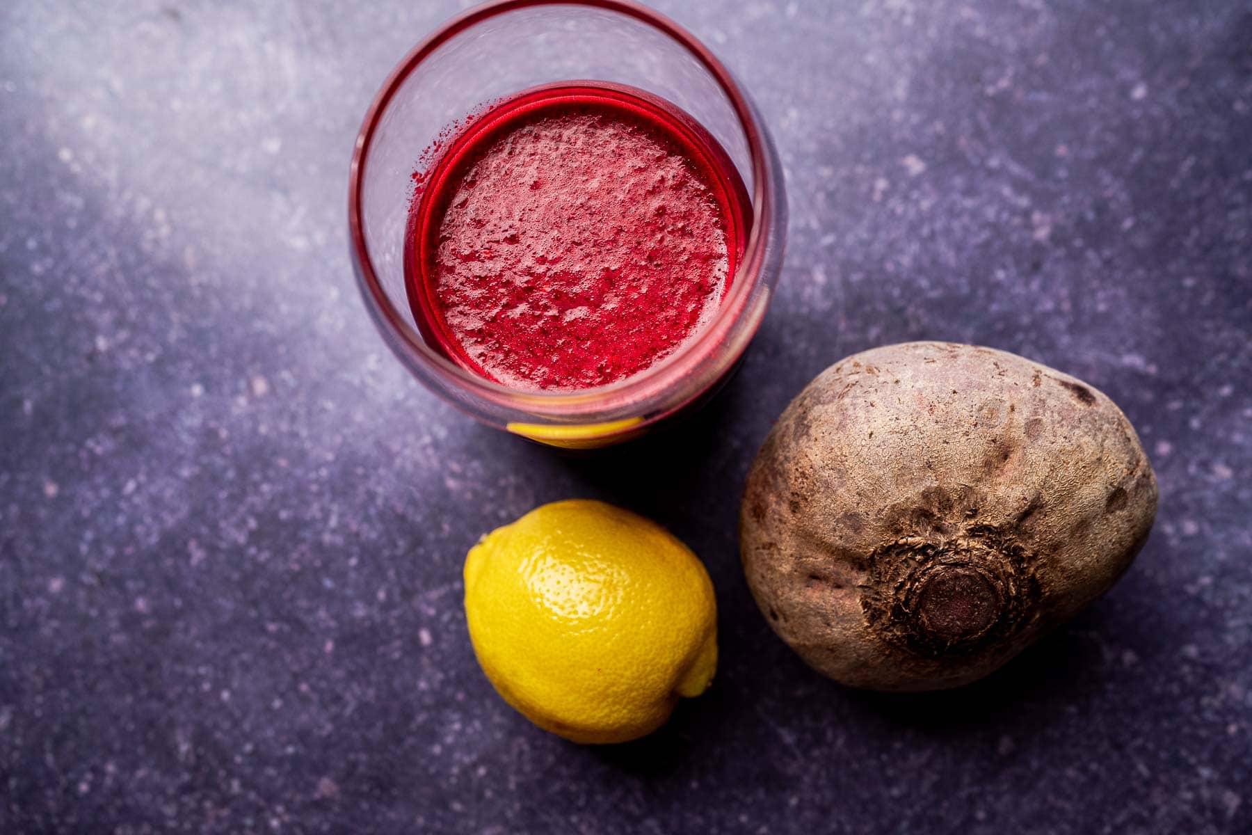 A glass of red beet pineapple juice sits on a dark textured surface next to a whole beetroot and a whole lemon.