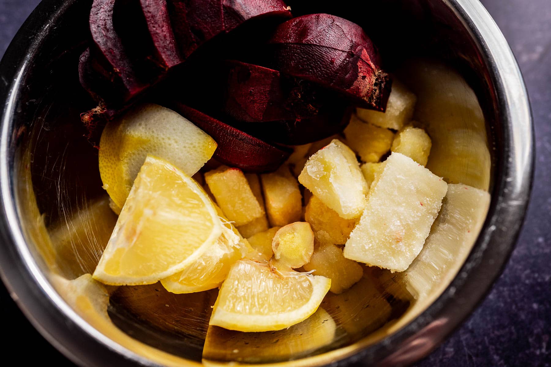 A metal bowl containing lemon wedges, frozen pineapple chunks, and dark red beet slices—perfect ingredients for a refreshing beet pineapple juice—rests on a dark surface.