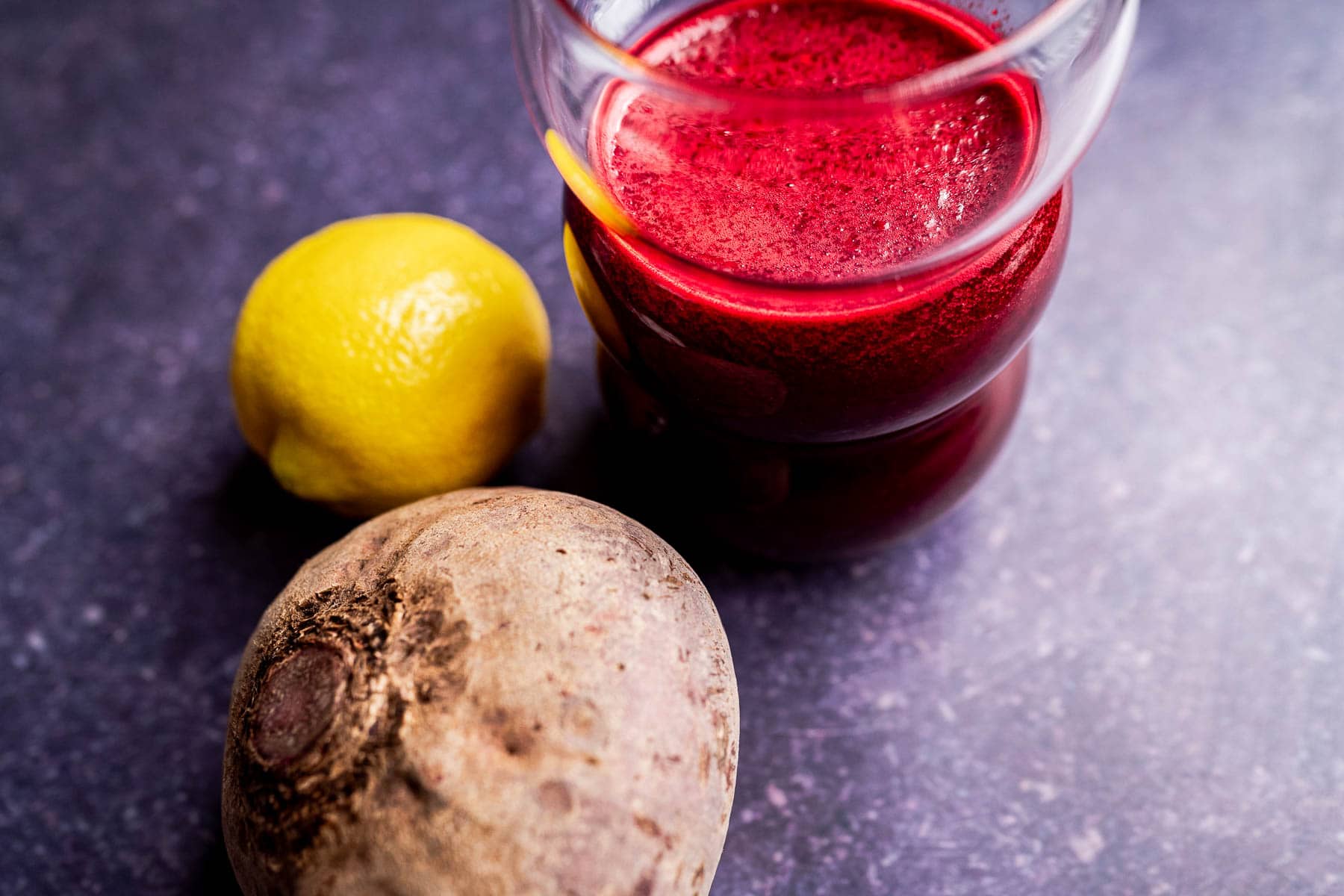 A glass of vibrant beet pineapple juice sits next to a whole beet and a lemon on a dark surface.