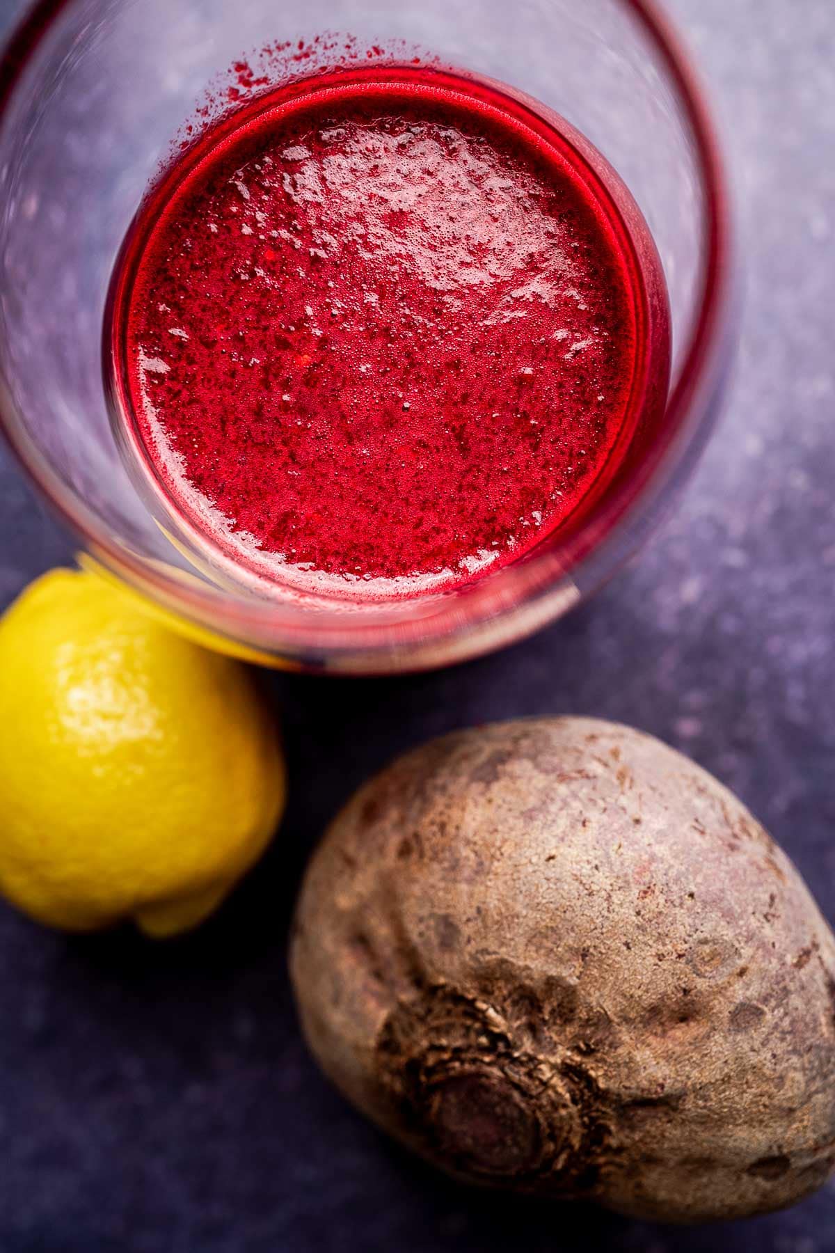 A glass of red beet pineapple juice on a dark surface with a whole beetroot and a lemon placed beside it.