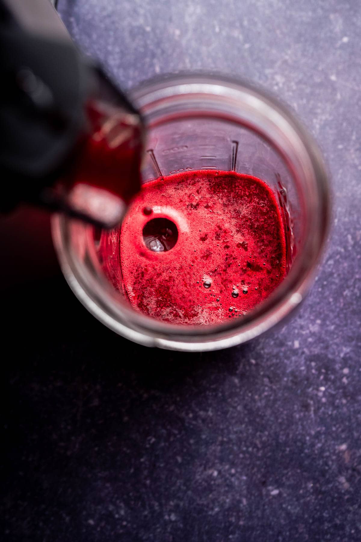 Overhead view of a blender cup containing a small amount of bright red beet pineapple juice on a dark surface.