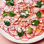 Thinly sliced radishes and red onions create a vibrant radish salad, topped with crumbled feta cheese, cilantro leaves, black pepper, and a drizzle of olive oil on a white plate.