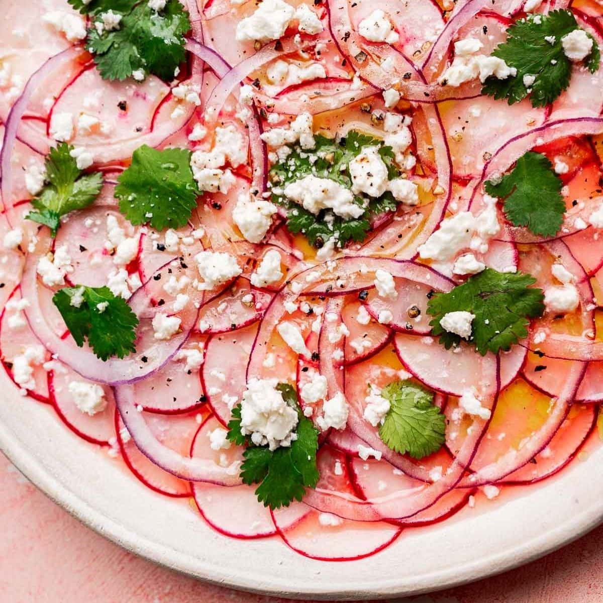 Thinly sliced radishes and red onions create a vibrant radish salad, topped with crumbled feta cheese, cilantro leaves, black pepper, and a drizzle of olive oil on a white plate.