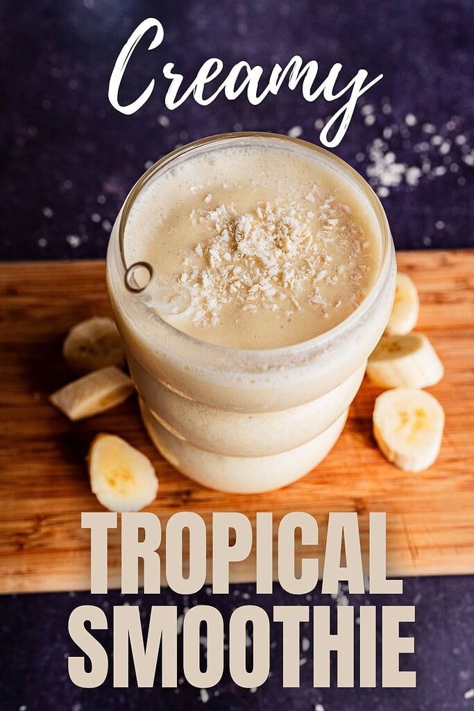 A creamy tropical smoothie topped with shredded coconut is served in a glass on a wooden board, surrounded by sliced bananas.