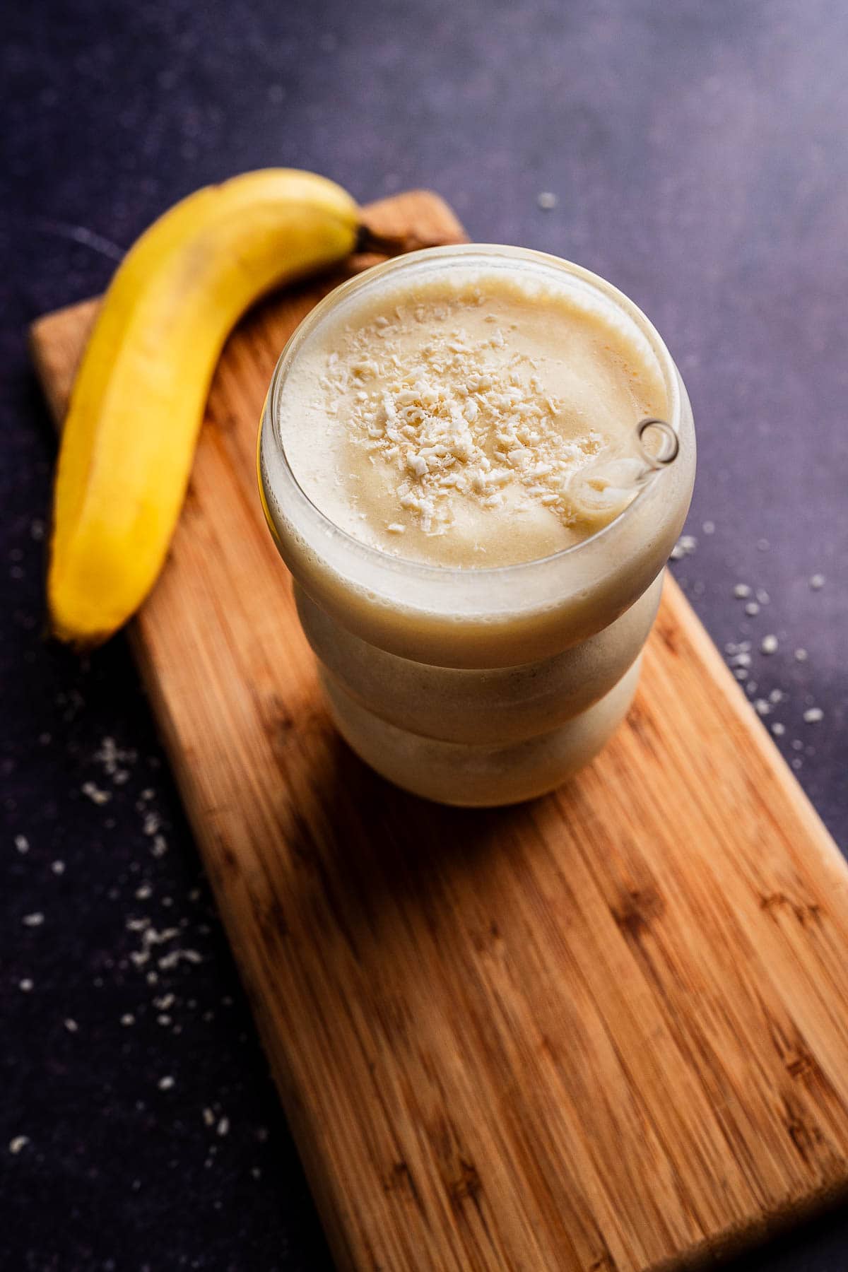 A glass of tropical smoothie, rich with banana and topped with shredded coconut, sits on a wooden board next to a whole banana.