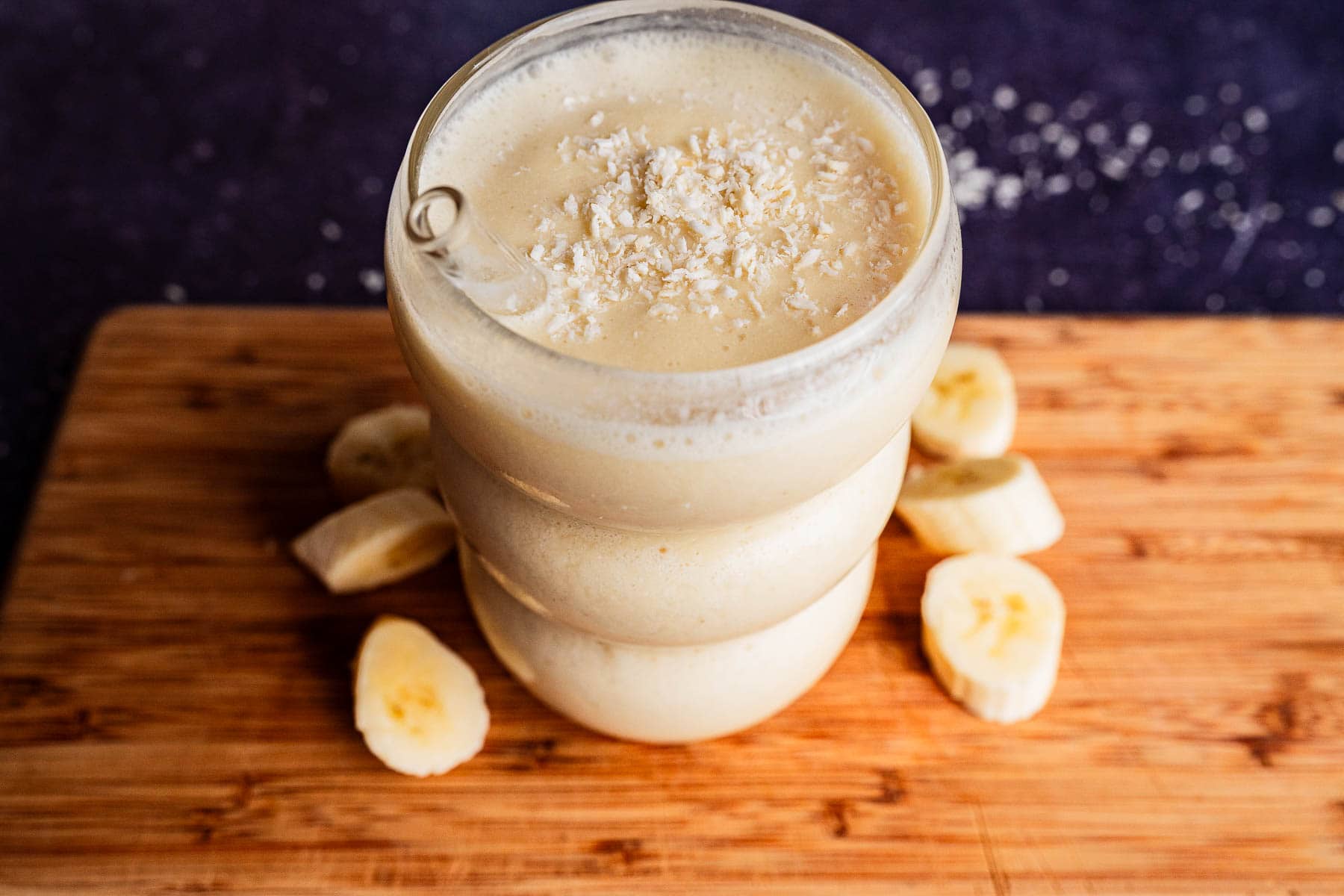 A glass of tropical smoothie topped with shredded coconut, placed on a wooden board with banana slices around it.