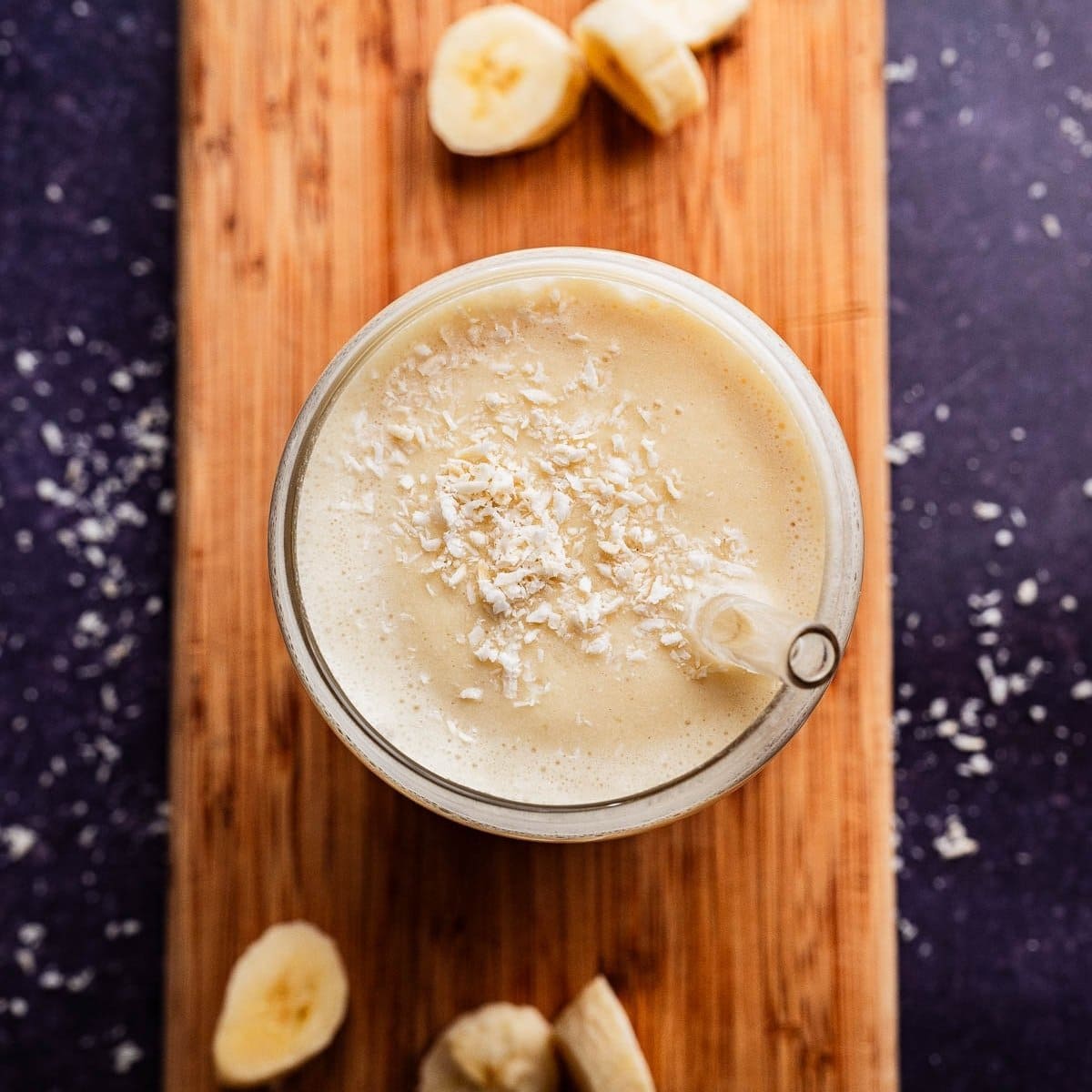 A glass of tropical smoothie topped with shredded coconut sits on a wooden board, with banana slices scattered around.