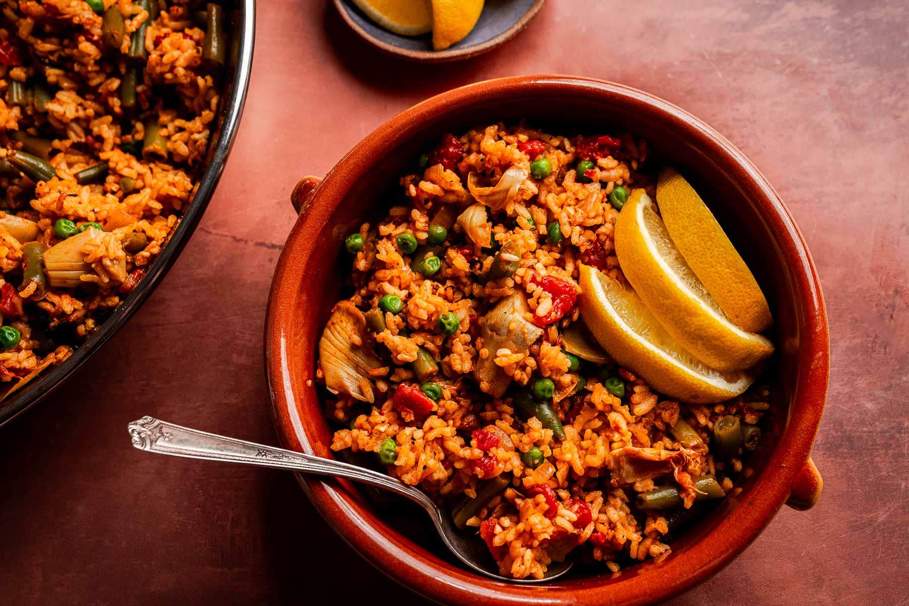 A bowl of Vegetarian Paella with peas, artichokes, tomatoes, and lemon wedges, served with a spoon on a brown surface.