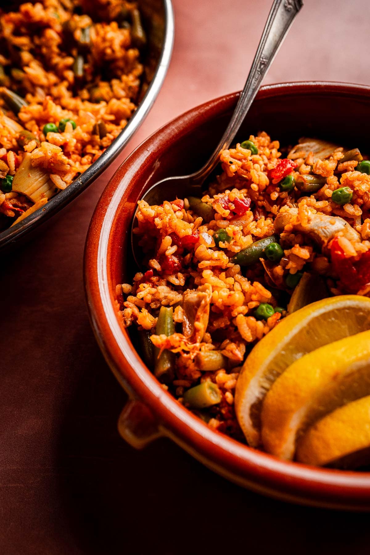 A bowl of Vegetarian Paella with peas, green beans, and lemon wedges, accompanied by a spoon and a serving dish in the background.