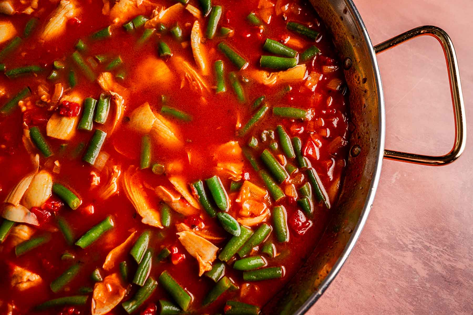 A pan filled with a tomato-based Vegetarian Paella, featuring tender artichoke hearts and green beans, sits on a light pink surface.