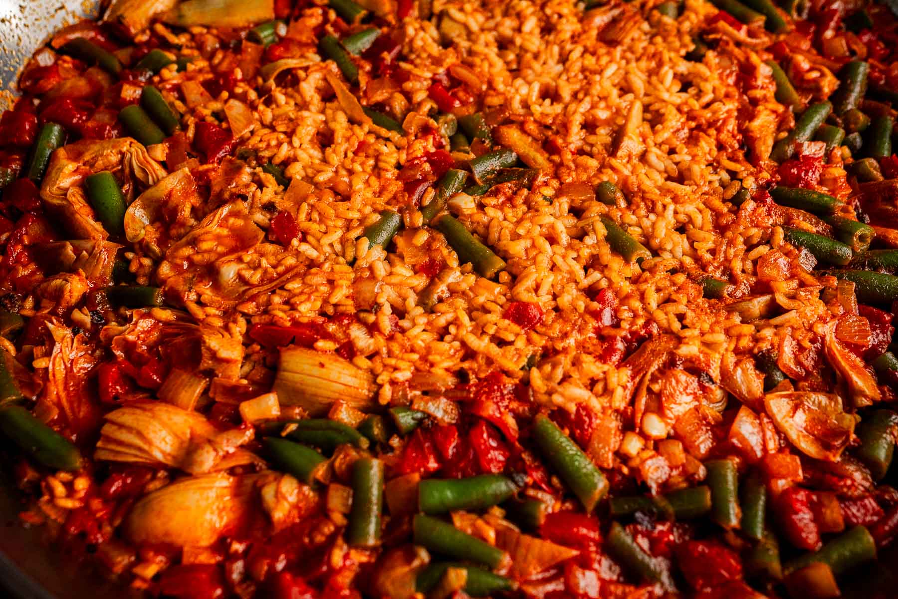 Close-up of a Vegetarian Paella featuring cooked rice, green beans, artichokes, and diced tomatoes in a pan.