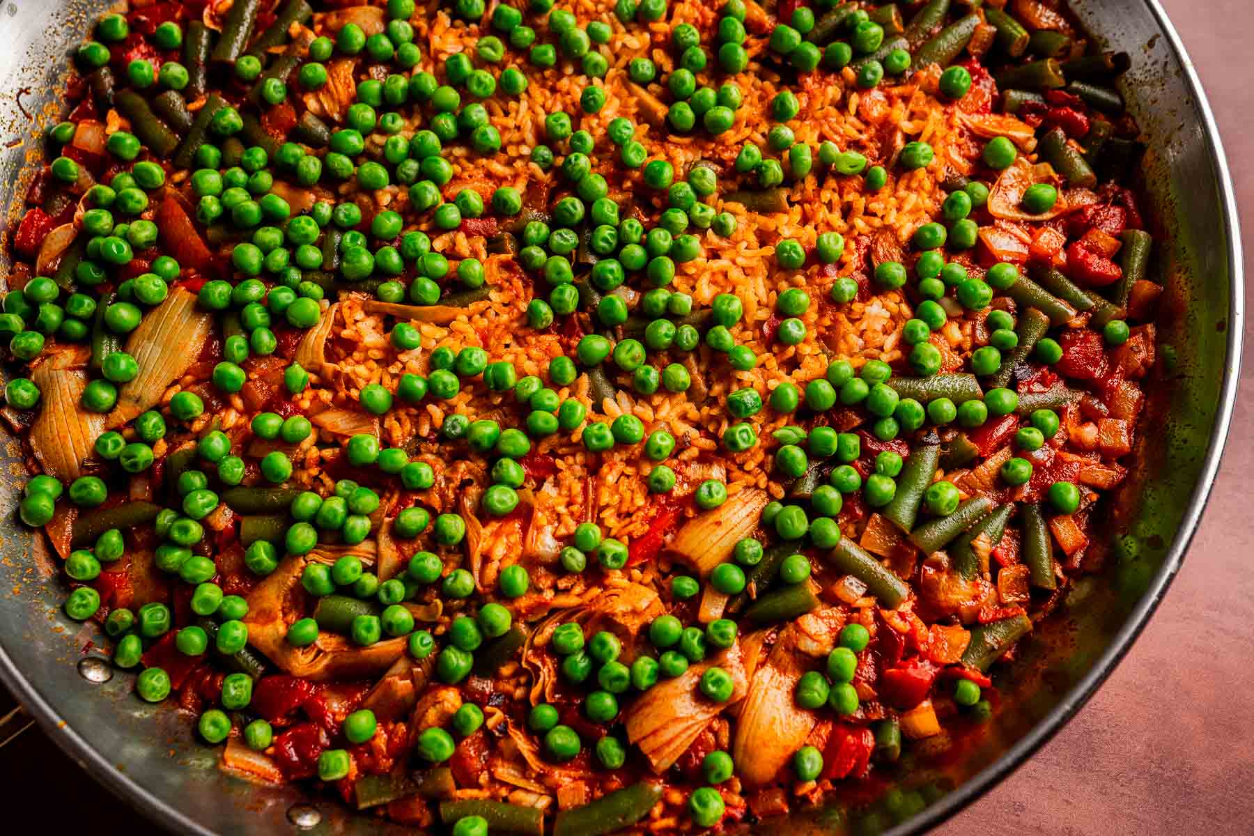 A close-up of a pan filled with Vegetarian Paella—rice, green peas, green beans, onions, and tomatoes all simmered together in a vibrant red sauce.
