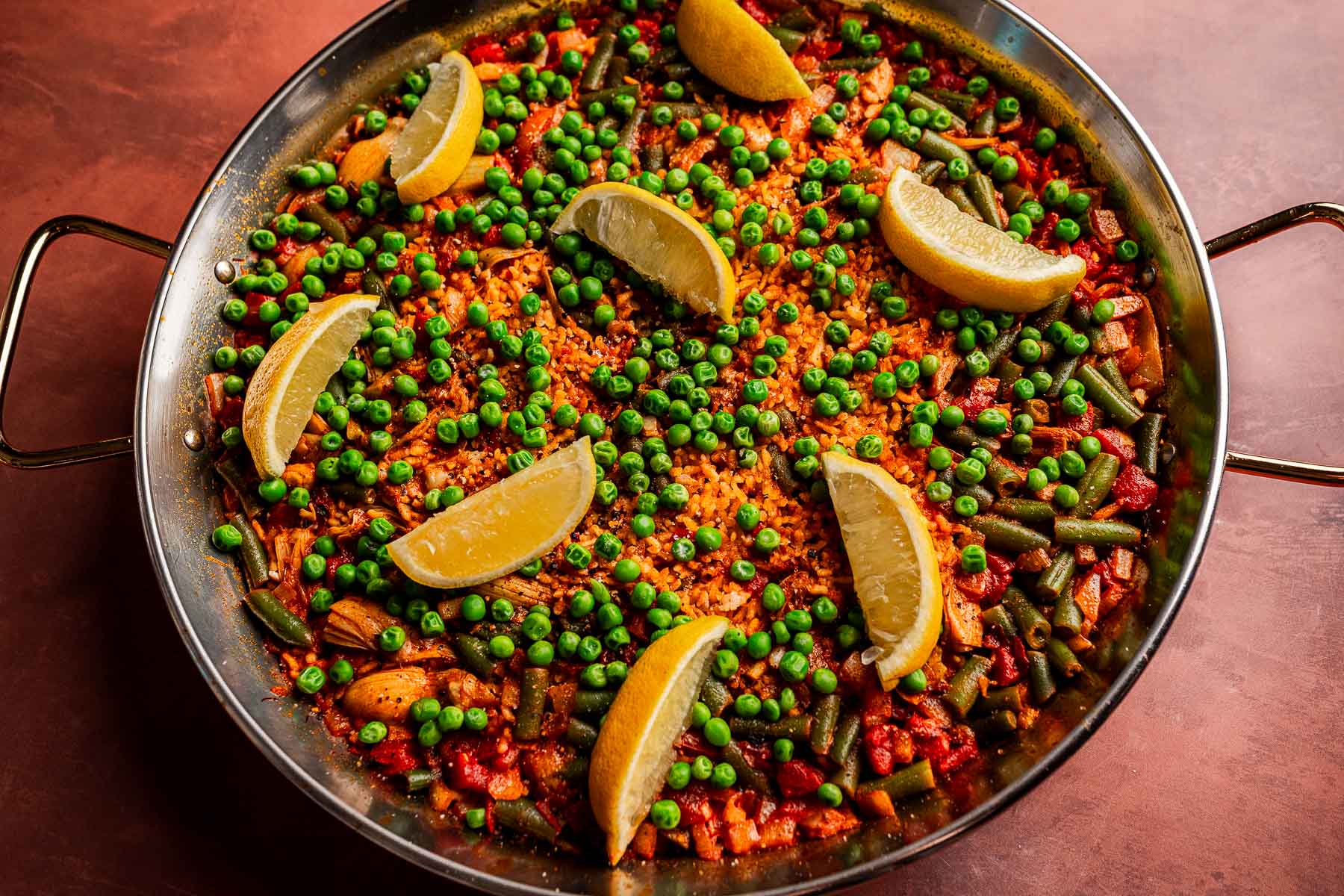 A pan of Vegetarian Paella topped with green peas and green beans, garnished with lemon wedges, sits on a brown surface.
