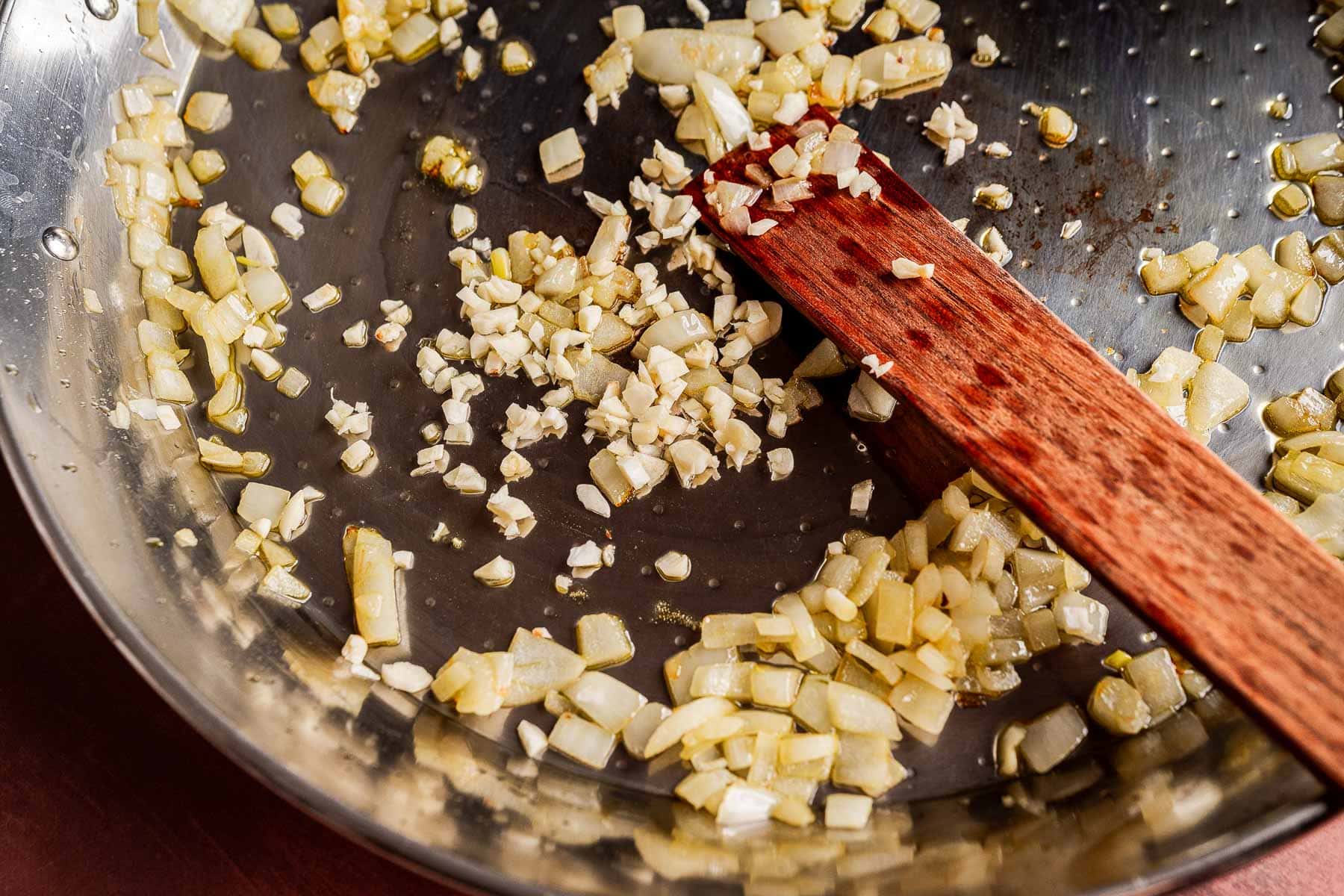 Chopped onions and garlic sauté in a stainless steel pan with a wooden spatula, forming the aromatic base for a delicious Vegetarian Paella.