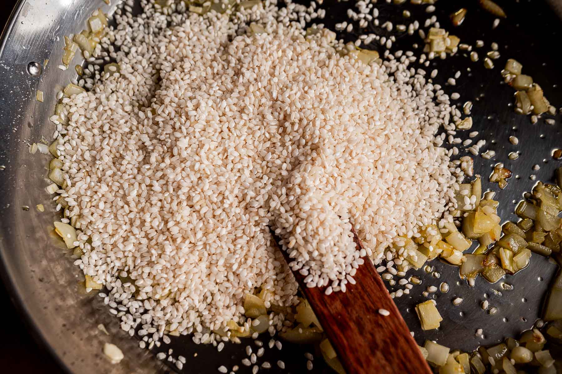 Uncooked arborio rice and chopped onions are being stirred in a metal pan with a wooden spatula, forming the base for a delicious Vegetarian Paella.