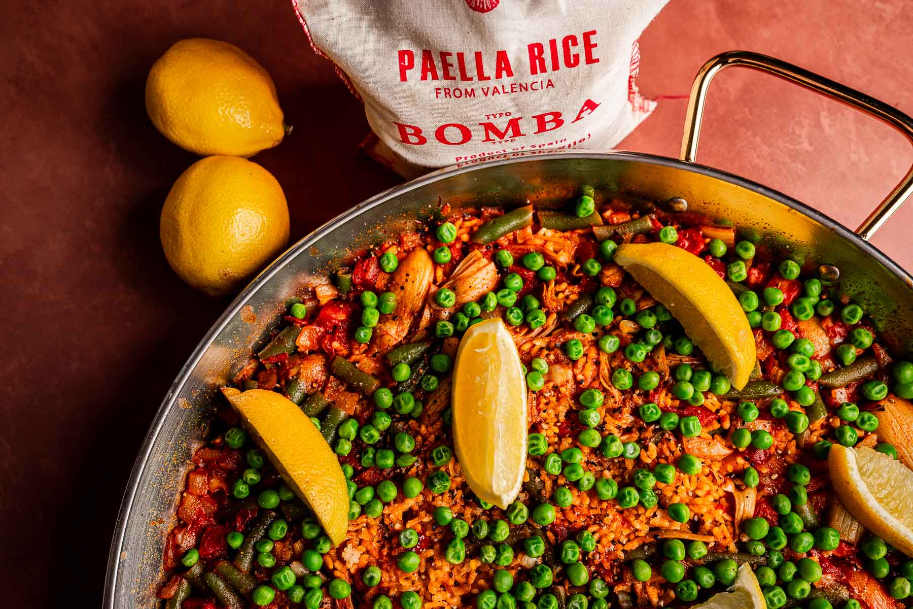 A pan of Vegetarian Paella with peas, lemon wedges, and vegetables sits beside two whole lemons and a bag labeled "Paella Rice from Valencia, Type Bomba" on a brown surface.