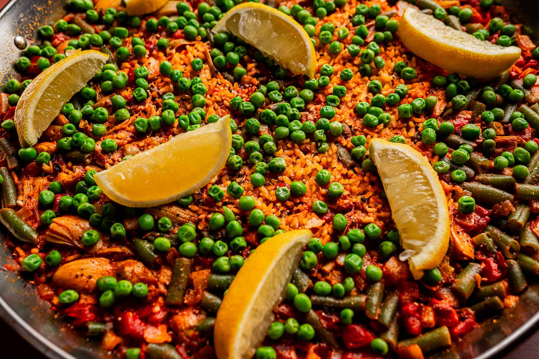 A close-up of a Vegetarian Paella, topped with green peas, green beans, and several lemon wedges on a bed of seasoned rice.