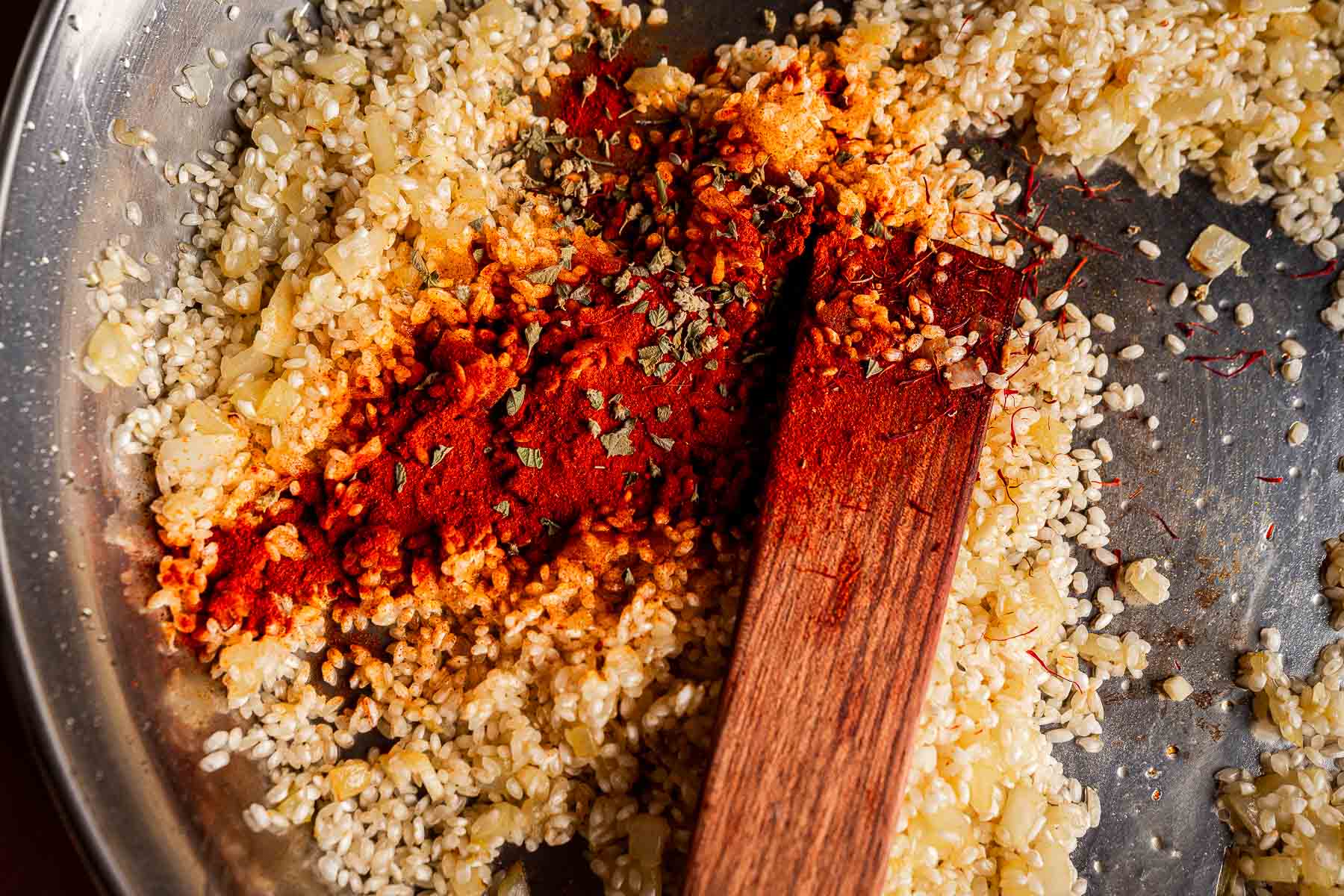 A close-up of a metal pan with sautéed onions, uncooked rice, paprika, herbs, and a wooden spatula mixing the ingredients—the perfect start for a flavorful Vegetarian Paella.