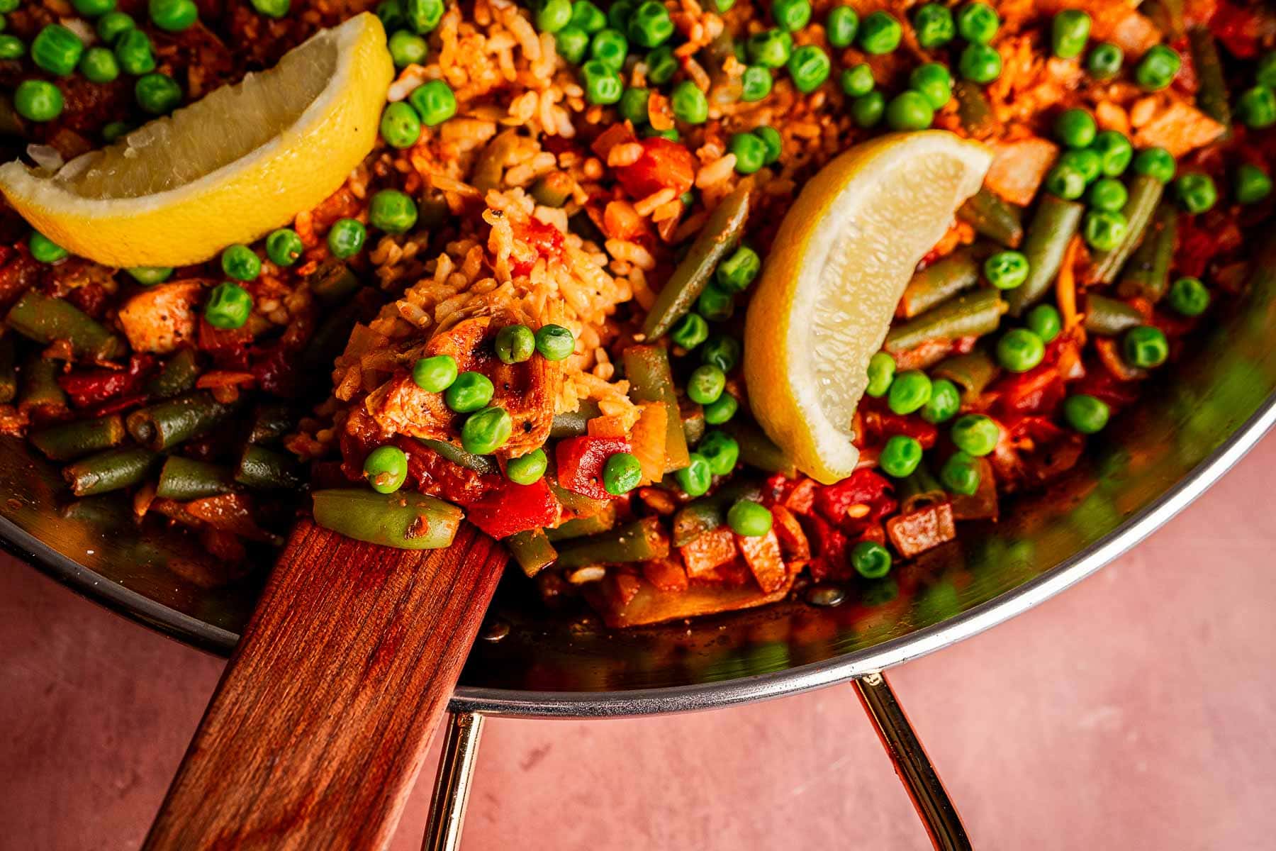A pan of Vegetarian Paella, featuring rice mixed with peas, green beans, diced vegetables, and tomato, garnished with two lemon wedges, as a wooden spatula serves a portion.