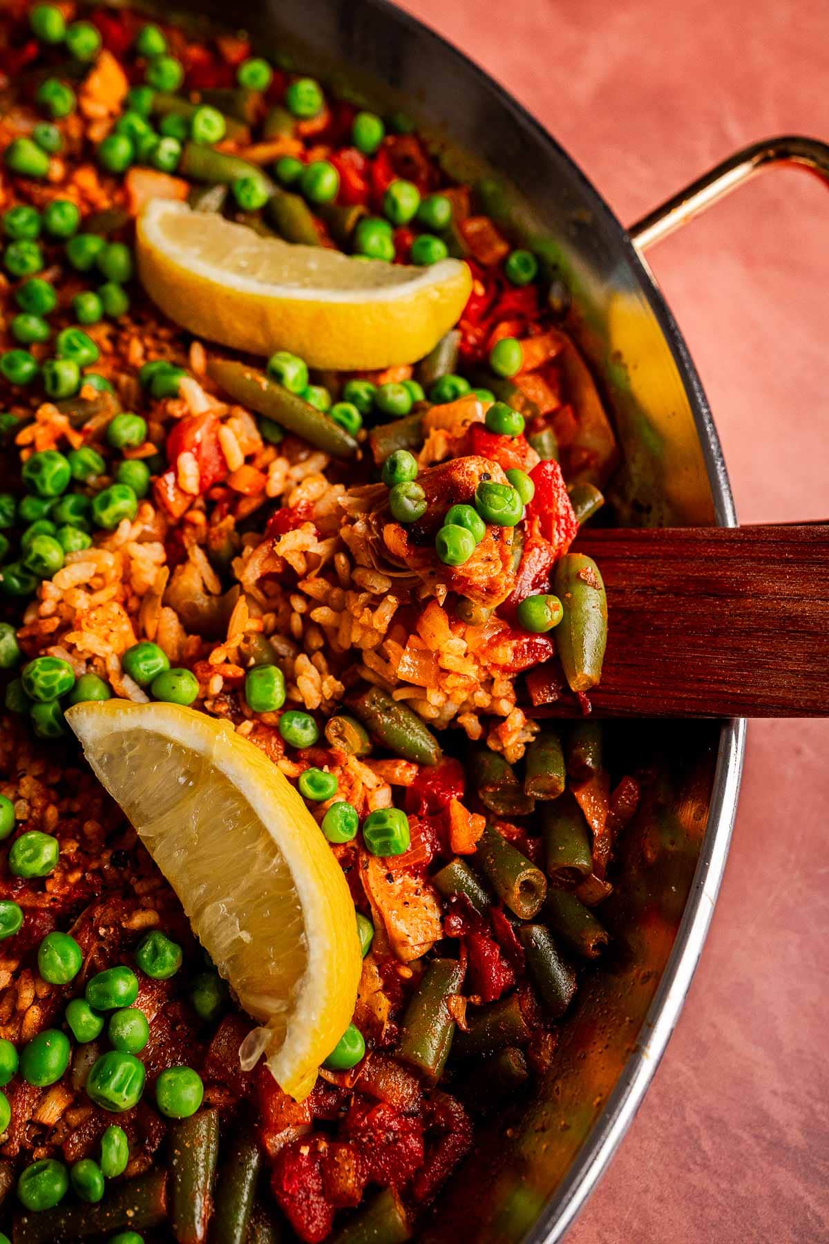 A pan of vibrant Vegetarian Paella, filled with rice and vegetables, is topped with green peas and lemon wedges, with a wooden spatula resting in the food.