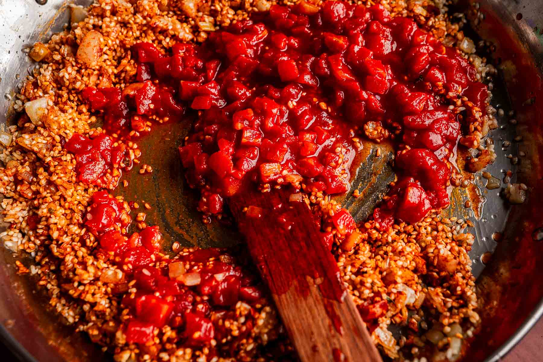 A close-up of a skillet with sautéed aromatics, uncooked bulgur, and diced tomatoes being stirred with a wooden spatula—the beginnings of a flavorful Vegetarian Paella.