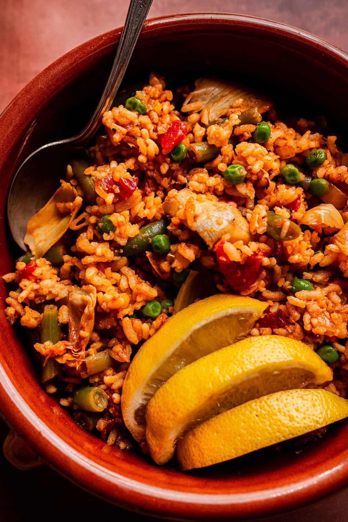 A bowl of Vegetarian Paella with peas, artichokes, and red peppers, garnished with three lemon wedges and served with a spoon.