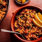A bowl of Vegetarian Paella with peas, artichokes, and red peppers, garnished with lemon wedges, sits beside a spoon and a plate holding extra lemon wedges.