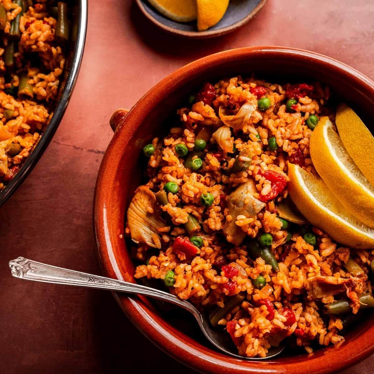 A bowl of Vegetarian Paella with peas, artichokes, and red peppers, garnished with lemon wedges, sits beside a spoon and a plate holding extra lemon wedges.