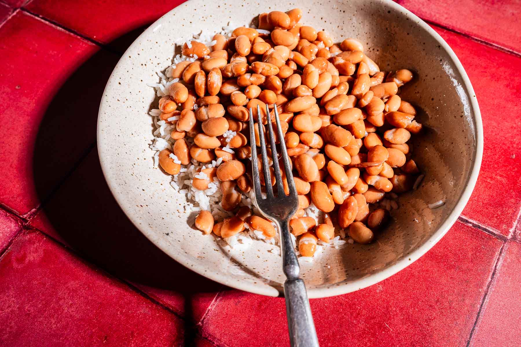 A bowl of white rice topped with pinto beans, with a metal fork resting inside, sits on a red tiled surface, reminiscent of the flavors found in a sheet pan crunchwrap.