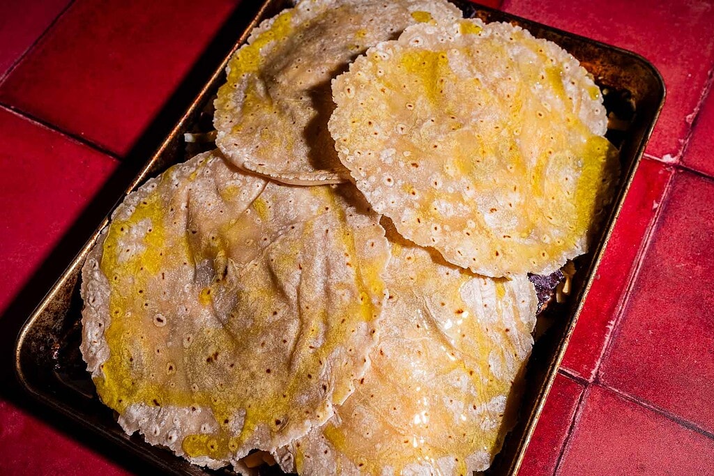 A metal tray holds a stack of yellow corn tortillas, perfect for making a homemade sheet pan crunchwrap, on a vibrant red tiled surface.