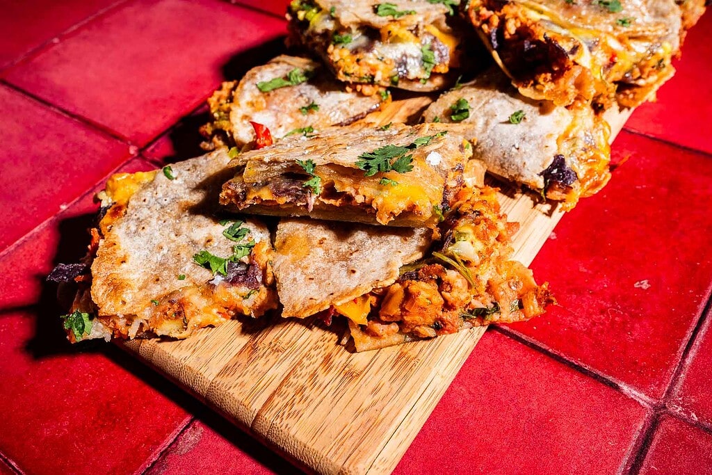 Sliced quesadillas filled with beans, vegetables, and cheese, served on a wooden board over a red tiled surface—like bite-sized sheet pan crunchwrap delights.