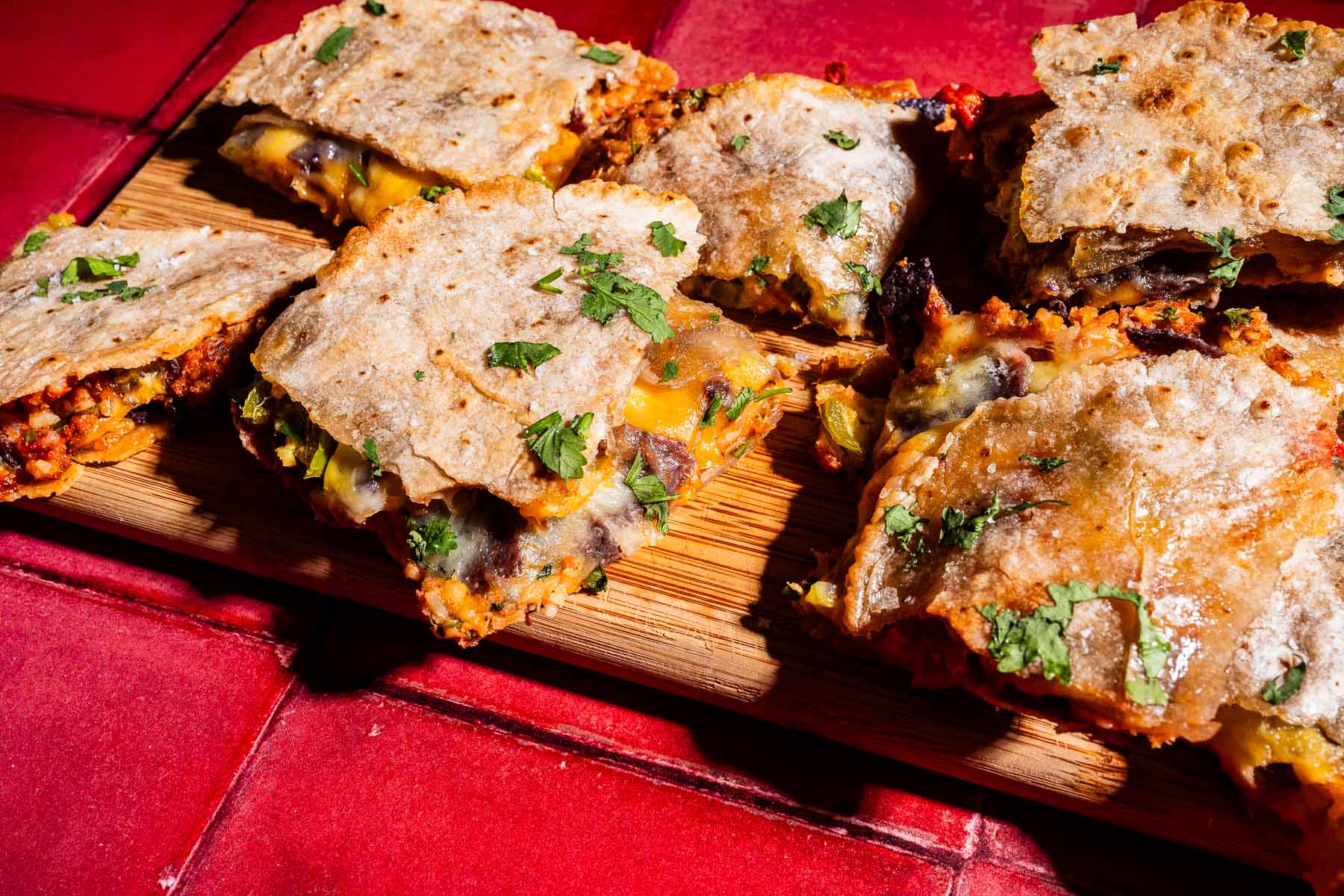 Six quesadillas filled with cheese and vegetables are arranged on a wooden board, garnished with chopped cilantro, on a red tiled surface—perfect for serving alongside your favorite sheet pan crunchwraps.
