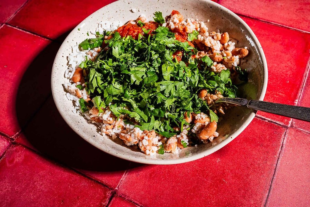 A bowl of rice and beans topped with chopped cilantro and red sauce, served alongside a crispy sheet pan crunchwrap, with a spoon, placed on a red tiled surface.