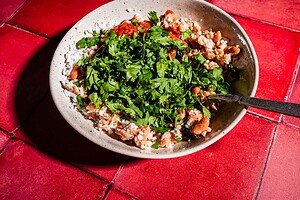 A bowl of rice and beans topped with chopped cilantro and red sauce, served alongside a crispy sheet pan crunchwrap, with a spoon, placed on a red tiled surface.