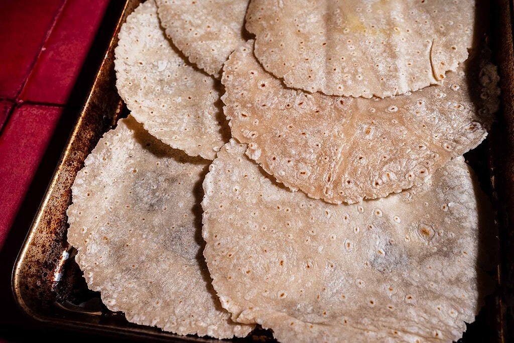 Several round, uncooked corn tortillas are stacked and overlapping on a metal baking sheet, ready to be transformed into a delicious sheet pan crunchwrap.