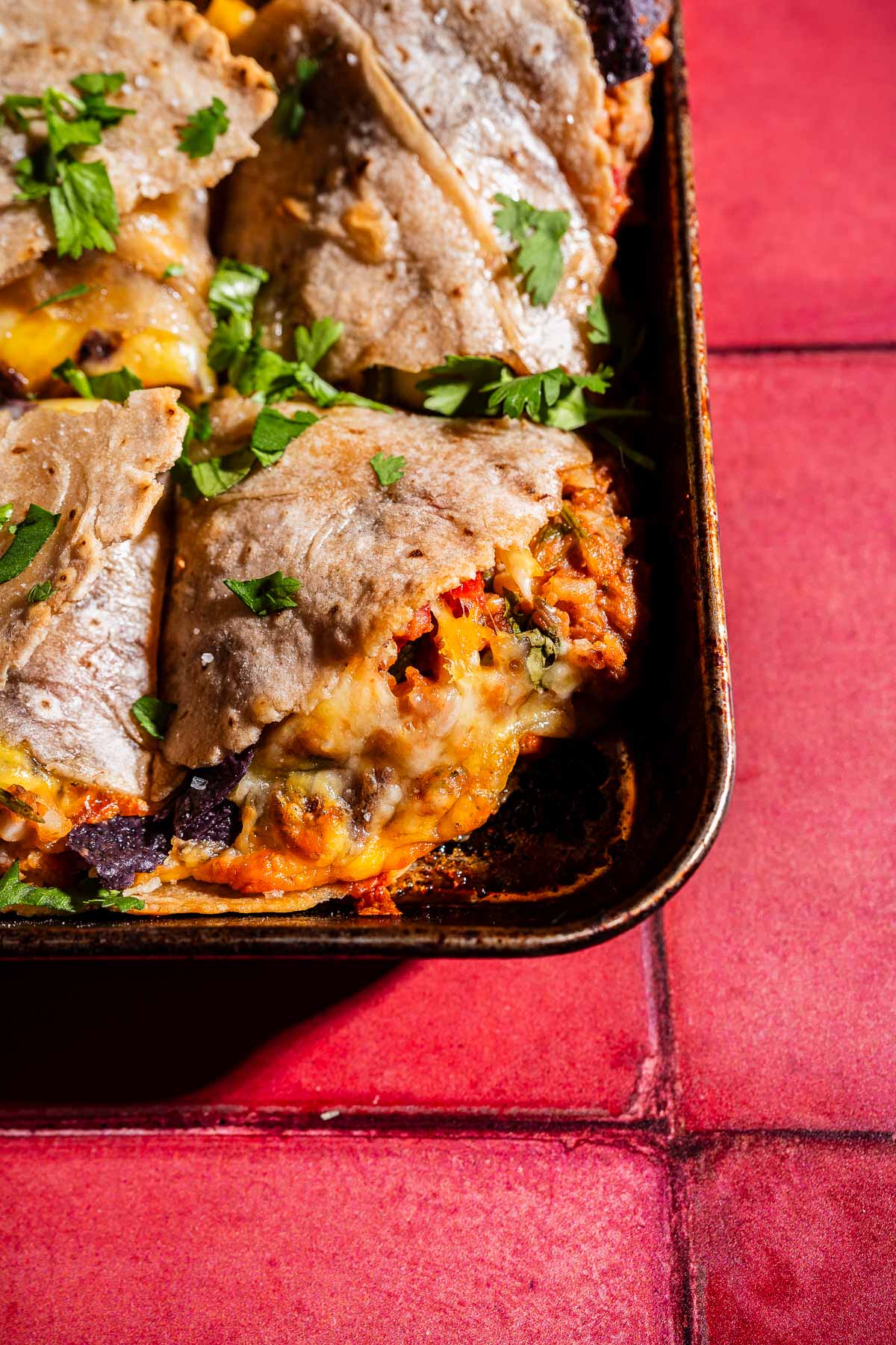 A close-up of baked enchiladas in a metal tray, topped with melted cheese and fresh cilantro, brings to mind the tempting layers of a sheet pan crunchwrap, all set on a vibrant red tiled surface.