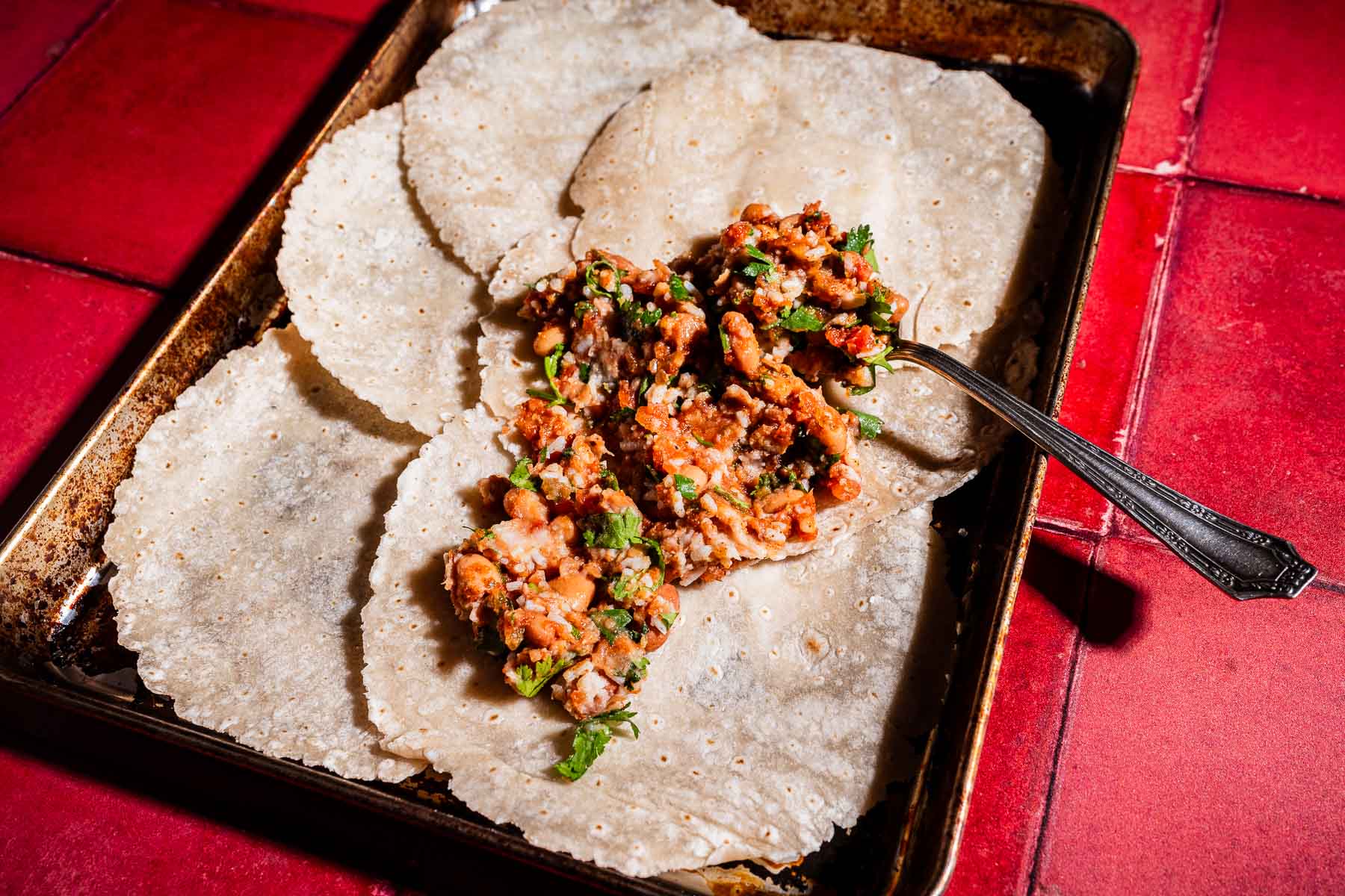 Four corn tortillas on a metal tray, with one topped like a sheet pan crunchwrap—filled with cooked meat, herbs, and vegetables. A spoon rests nearby on the red tiled surface underneath.