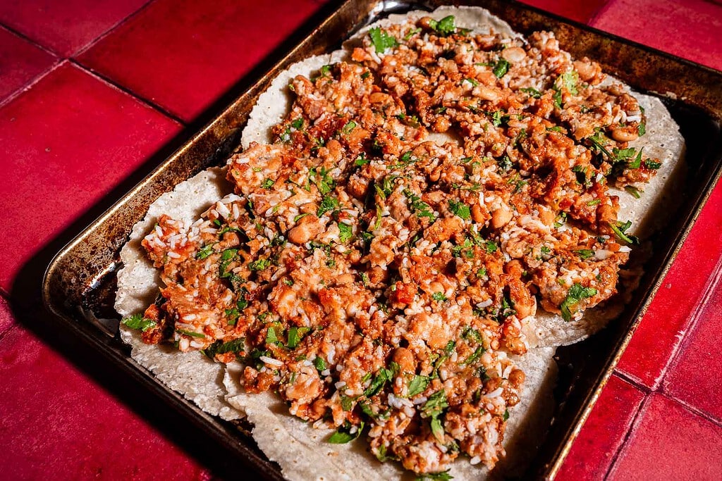 A baking sheet with a large, flat tortilla topped with cooked ground meat, beans, herbs, and sauce—perfect for making delicious sheet pan crunchwrap quesadillas—placed on a red tiled surface.