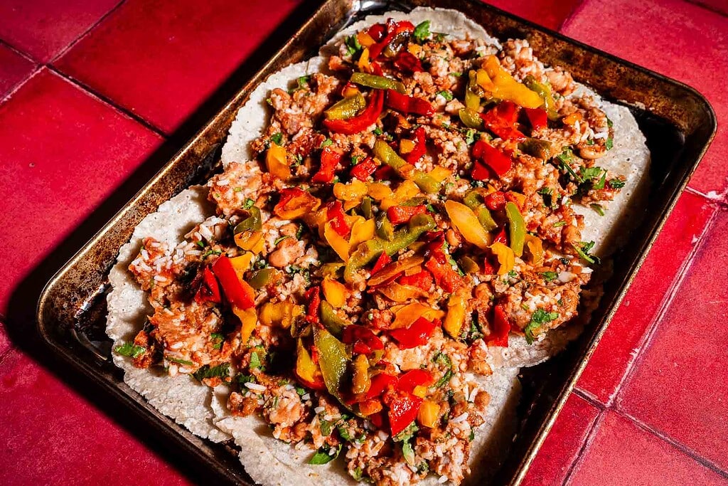 A baking tray on a red tiled surface holds a flat dish, reminiscent of a sheet pan crunchwrap, topped with rice, beans, and sliced red, yellow, and green peppers.