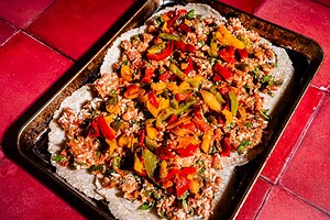 A baking tray on a red tiled surface holds a flat dish, reminiscent of a sheet pan crunchwrap, topped with rice, beans, and sliced red, yellow, and green peppers.